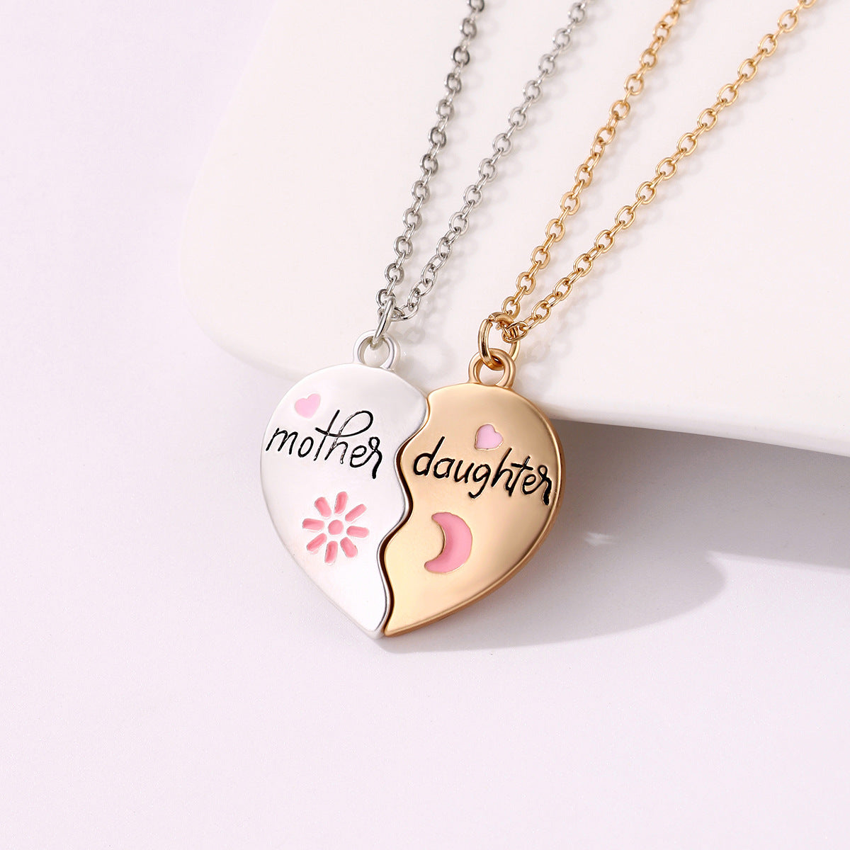 A two-piece heart-shaped necklace set with "Mother" on a silver pendant and "Daughter" on a gold pendant, featuring pink heart and moon details.