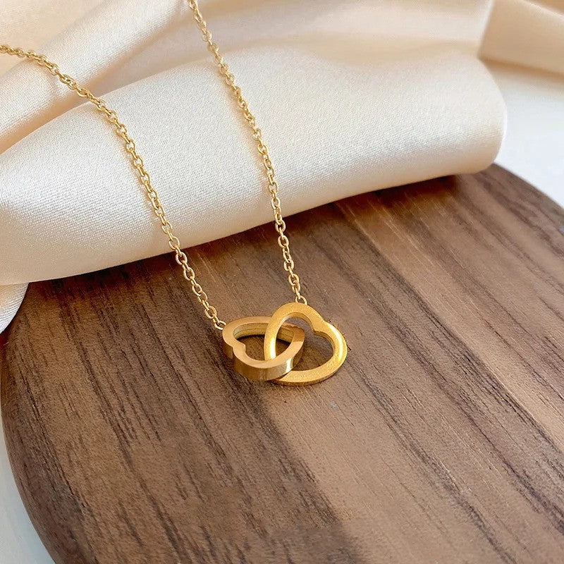 Close-up of the Solo Love Interlocking Hearts Necklace in gold stainless steel, featuring two intertwined heart pendants displayed on a table