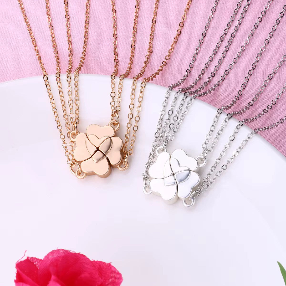 Both gold and silver Four-Leaf Clover Heart Magnetic Necklaces displayed side by side on soft pink fabric and a white plate. Each pendant features an interlocking heart design, symbolizing connection and love, with delicate chains.